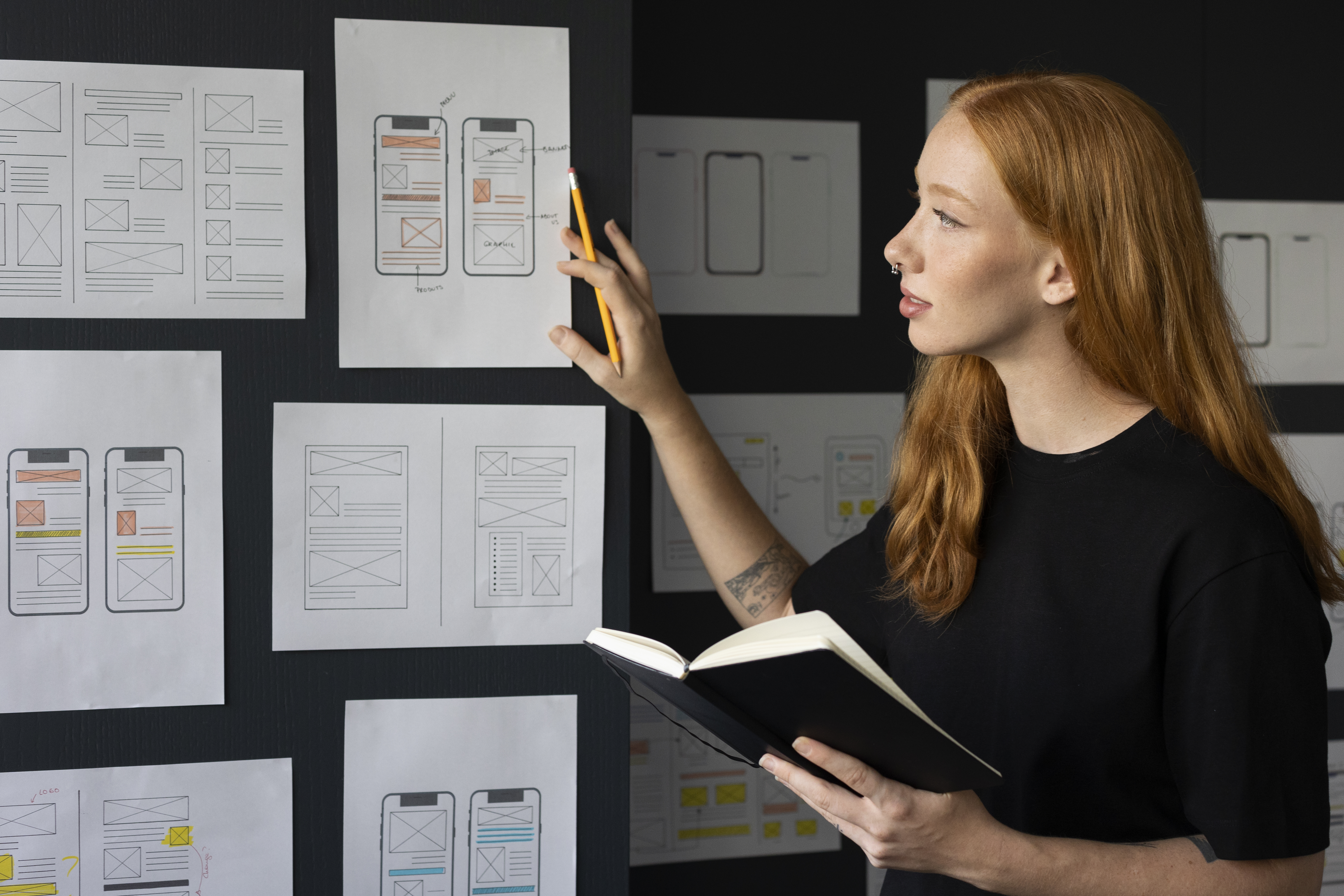 A mobile app designer points to paper wireframes of phone screens displayed on a wall while reviewing sketches in a notebook.