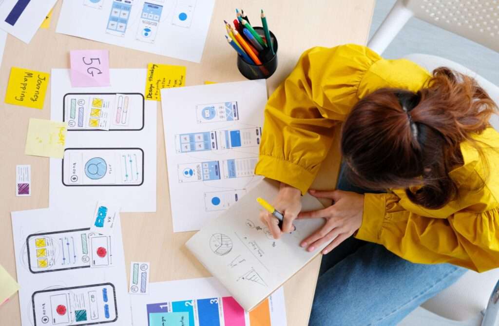 A designer in a yellow jacket sketches ideas beside printed mobile interface wireframes and smartphone screen layouts on a desk.