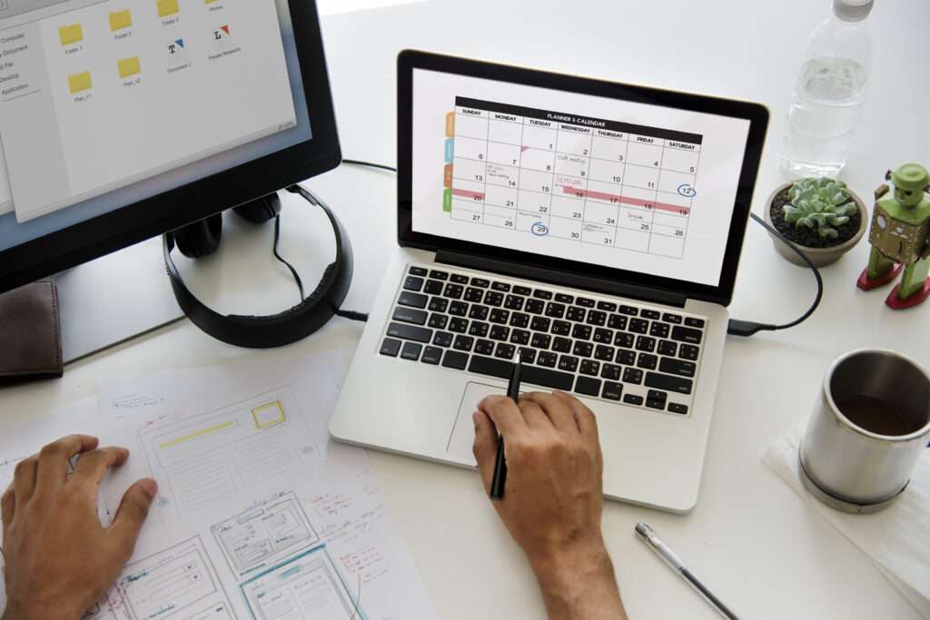 A laptop displays a calendar beside a desktop monitor and project sketches on a desk, illustrating development scheduling and milestone planning.